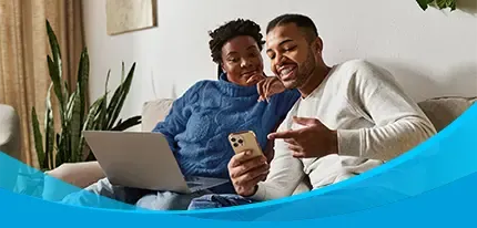 A young couple sits together on a sofa, looking at a smartphone while a laptop rests on one person's lap.